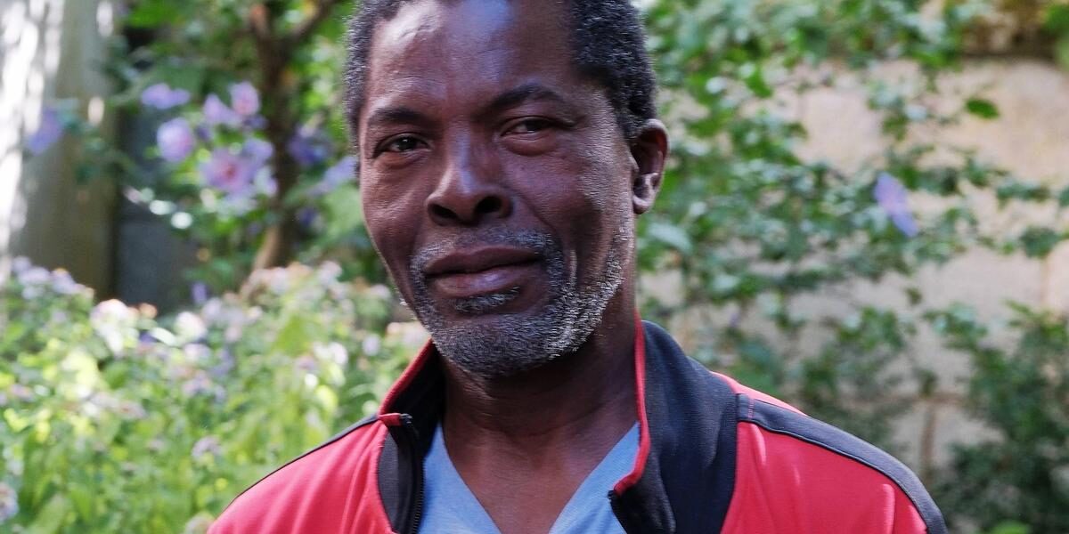 Close-up of a smiling Black man wearing a red jacket, outdoors in a garden setting.