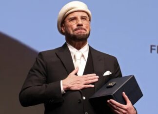 Man in a black suit and white beret holds a dark rectangular box on stage, hand over his chest.