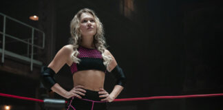 Athlete in a wrestling ring poses with hands on hips, wearing a pink-and-black sports bra and shorts and elbow guards.