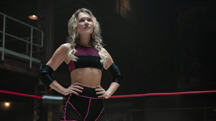 Athlete in a wrestling ring poses with hands on hips, wearing a pink-and-black sports bra and shorts and elbow guards.