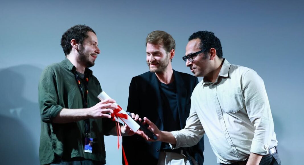 Laser Gato - Lucas Acher Cannes 2026 Three men on a stage exchange a rolled certificate tied with a red ribbon, as one presenter smiles at another.
