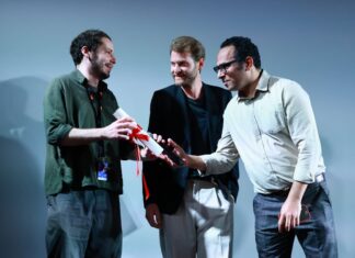 Cannes 2026 | Brasileiro Lucas Acher vence La Cinef com ‘Laser-Gato’, estrelado por Gilda Nomacce Three men on a stage exchange a rolled certificate tied with a red ribbon, as one presenter smiles at another.