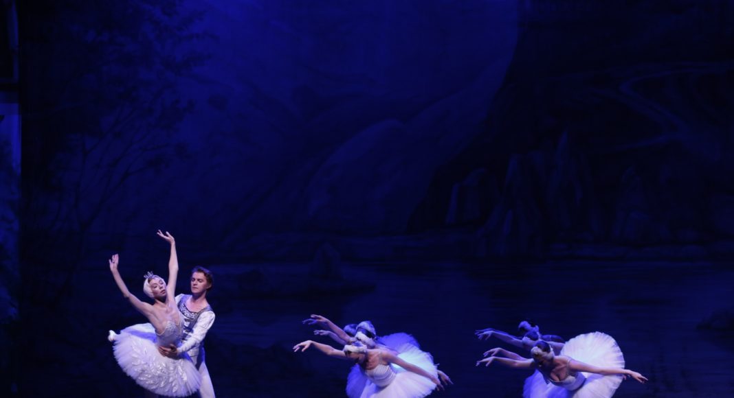 Ballet couple performing a lift on a blue-lit stage, with several white-tutu dancers in the background.