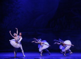 Ballet couple performing a lift on a blue-lit stage, with several white-tutu dancers in the background.