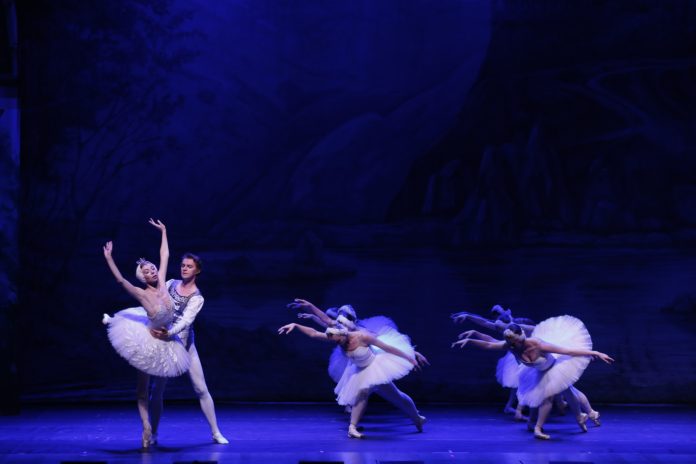 Ballet couple performing a lift on a blue-lit stage, with several white-tutu dancers in the background.