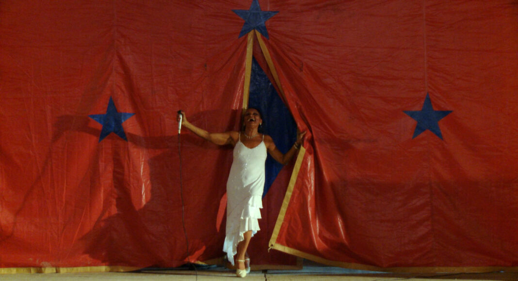 Female performer in a white dress singing into a microphone on a stage with a red backdrop and blue stars behind her.