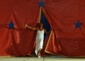 Female performer in a white dress singing into a microphone on a stage with a red backdrop and blue stars behind her.
