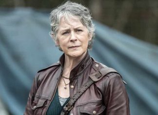 Woman with short gray hair in a brown leather jacket, standing outdoors with a blue tent in the background.