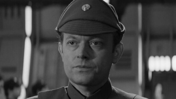 Black-and-white portrait of a man in a vintage military uniform and cap, looking slightly to the left in a blurred indoor setting.
