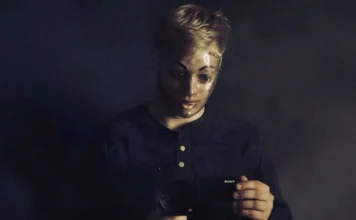 Self-portrait of a person in a dark room holding a camera, with facial makeup and a small 'MOLD' mark on the forehead visible in the reflection/face paint.