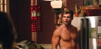 Muscular, shirtless man stands in a cluttered kitchen beside a stainless steel refrigerator, looking off to the side with a serious expression.