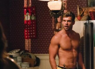 Muscular, shirtless man stands in a cluttered kitchen beside a stainless steel refrigerator, looking off to the side with a serious expression.