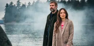 A bearded man in a dark coat and a woman in a beige blazer stand near a misty lake with trees in the background.