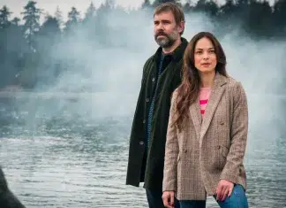 A bearded man in a dark coat and a woman in a beige blazer stand near a misty lake with trees in the background.