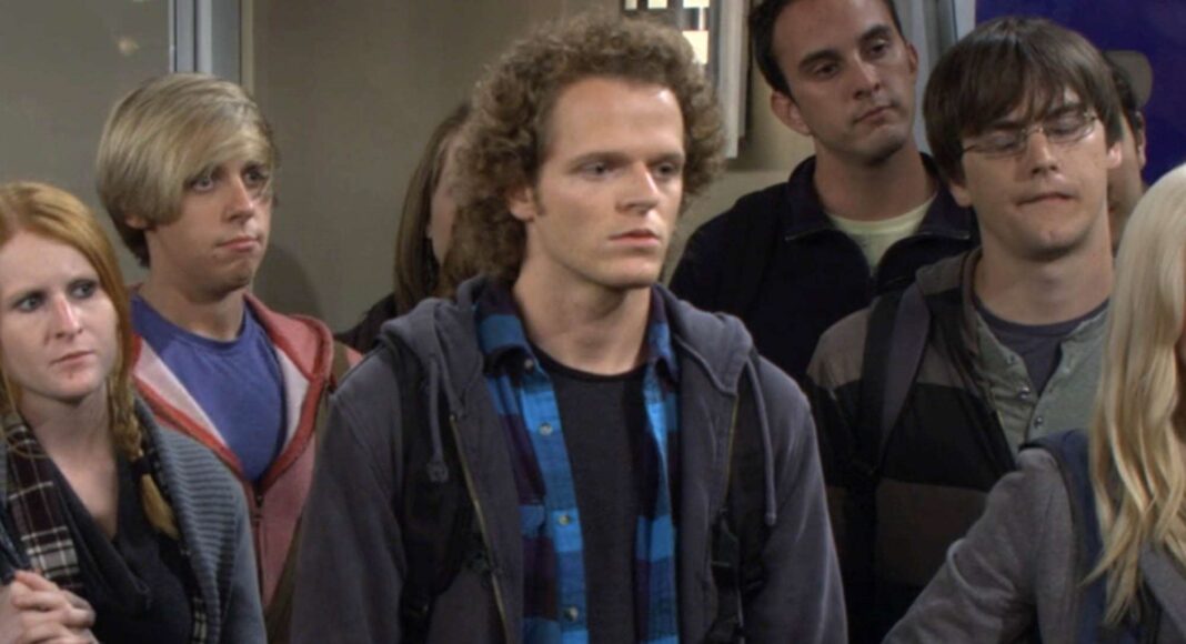 Nick-Pasqual-2 Group of young adults standing indoors, looking ahead with neutral expressions; center man with curly hair in a blue plaid shirt.