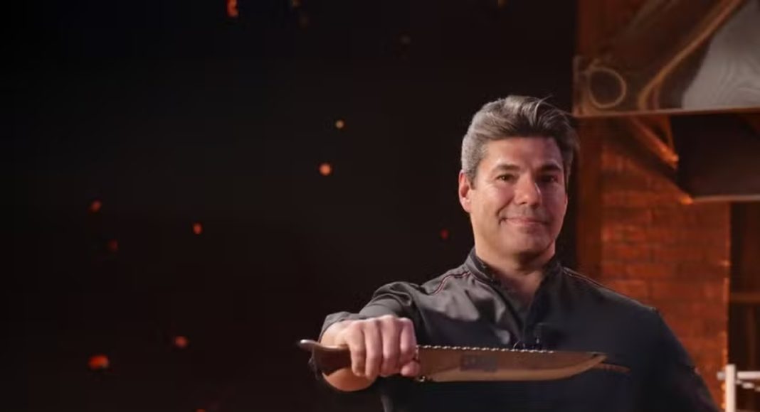 Chef in black uniform extending a large knife toward the camera in a studio kitchen backdrop with brick walls and warm lighting.