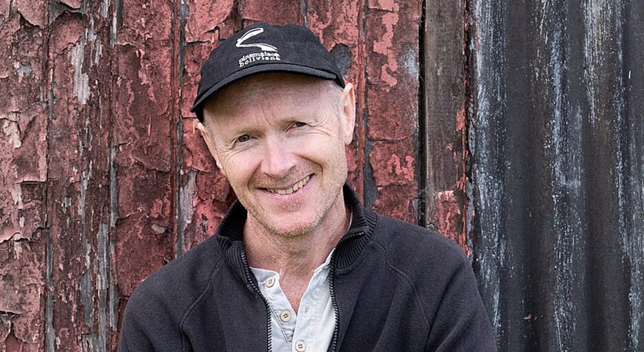 Smiling man with arms crossed in front of weathered red and black wooden planks, wearing a black zip-up jacket and cap.