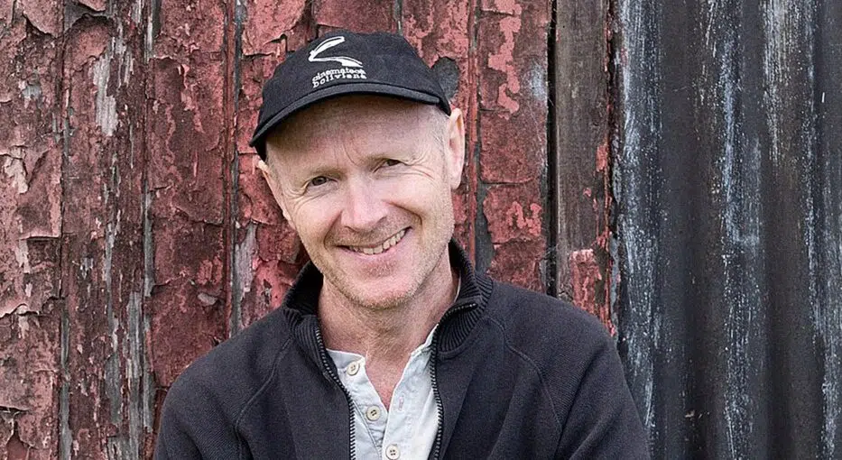 Smiling man with arms crossed in front of weathered red and black wooden planks, wearing a black zip-up jacket and cap.