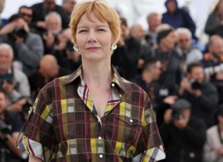 Woman with short reddish hair in a plaid shirt at a public event, with photographers in the background.