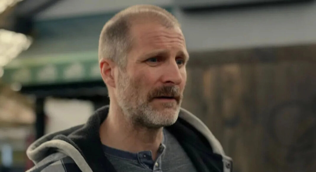 Middle-aged man with short gray beard in a gray henley and hoodie, indoors in a workshop or warehouse setting.