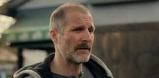 Middle-aged man with short gray beard in a gray henley and hoodie, indoors in a workshop or warehouse setting.