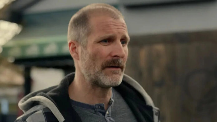 Middle-aged man with short gray beard in a gray henley and hoodie, indoors in a workshop or warehouse setting.