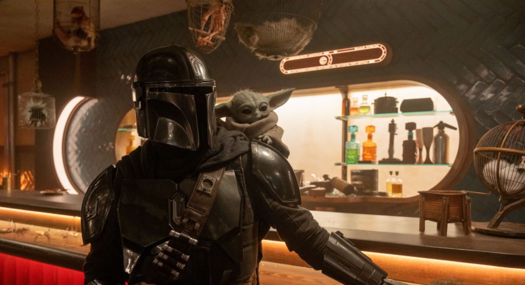 Mandalorian in black armor at a bar with Grogu perched on his shoulder, dim sci‑fi tavern setting.
