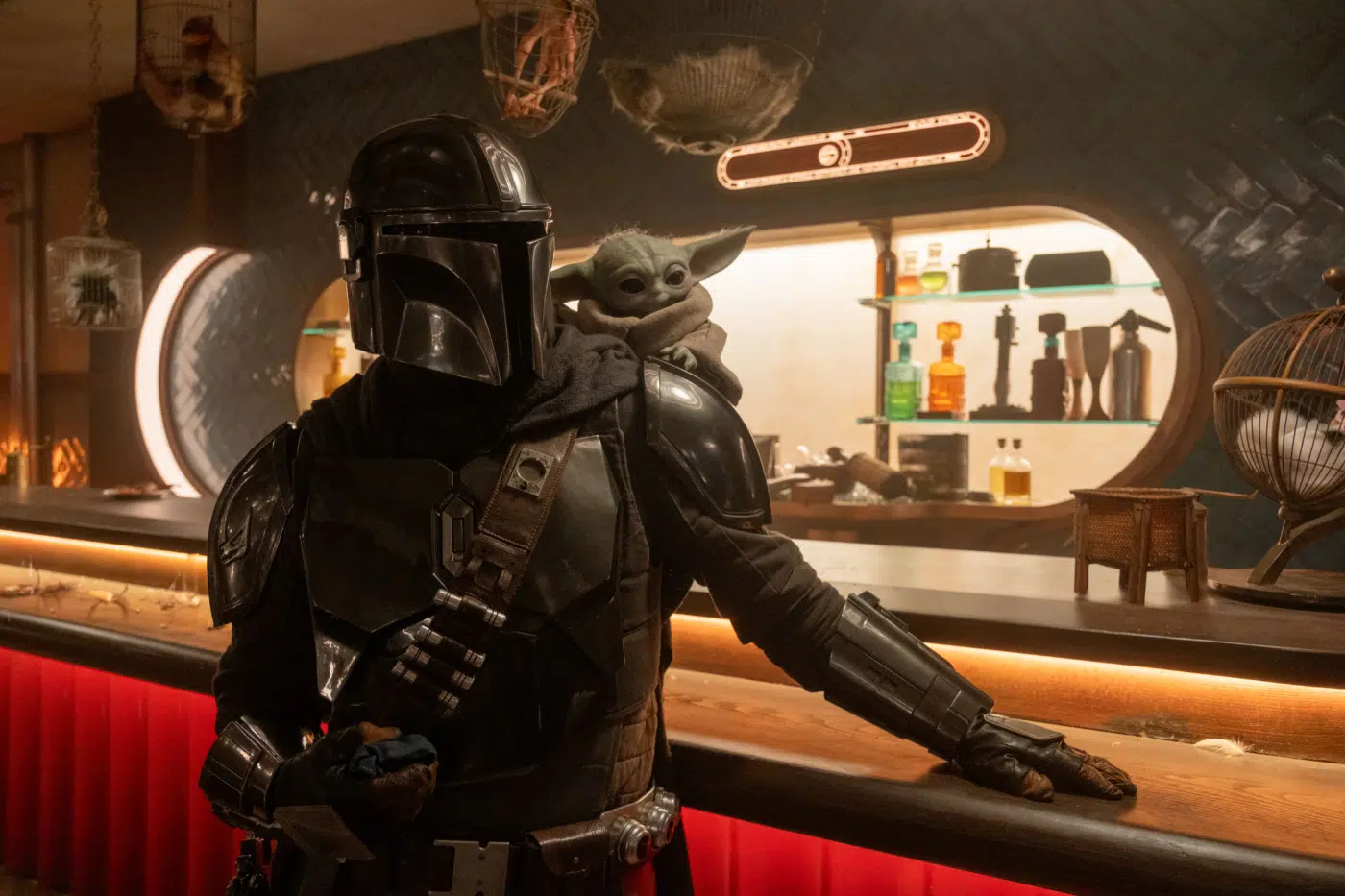 Mandalorian in black armor at a bar with Grogu perched on his shoulder, dim sci‑fi tavern setting.