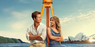 Couple sitting back-to-back on a yellow boat frame, arms crossed, smiling at each other with Sydney Opera House in the background.