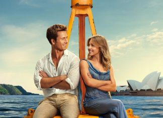 Dicas de filmes para o fim de semana | Comédias românticas APAIXONANTES para curtir nos streamings Couple sitting back-to-back on a yellow boat frame, arms crossed, smiling at each other with Sydney Opera House in the background.