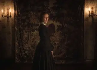 Com Maika Monroe, terror GÓTICO ‘Psicopata Vitoriana’ ganha data de estreia nos cinemas nacionais Woman in 19th-century dress stands in a dimly lit room with a tapestry backdrop and wall sconces.