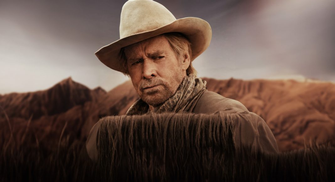 Man in a beige cowboy hat and scarf stands behind tall dry grass in a desert landscape with hills in the background, looking thoughtful.