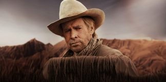 Man in a beige cowboy hat and scarf stands behind tall dry grass in a desert landscape with hills in the background, looking thoughtful.