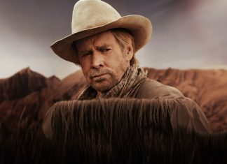 Man in a beige cowboy hat and scarf stands behind tall dry grass in a desert landscape with hills in the background, looking thoughtful.