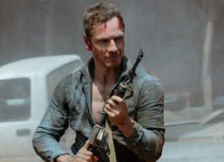 Intense man with a bloodied forehead holding a rifle in a dusty, ruined outdoor scene.