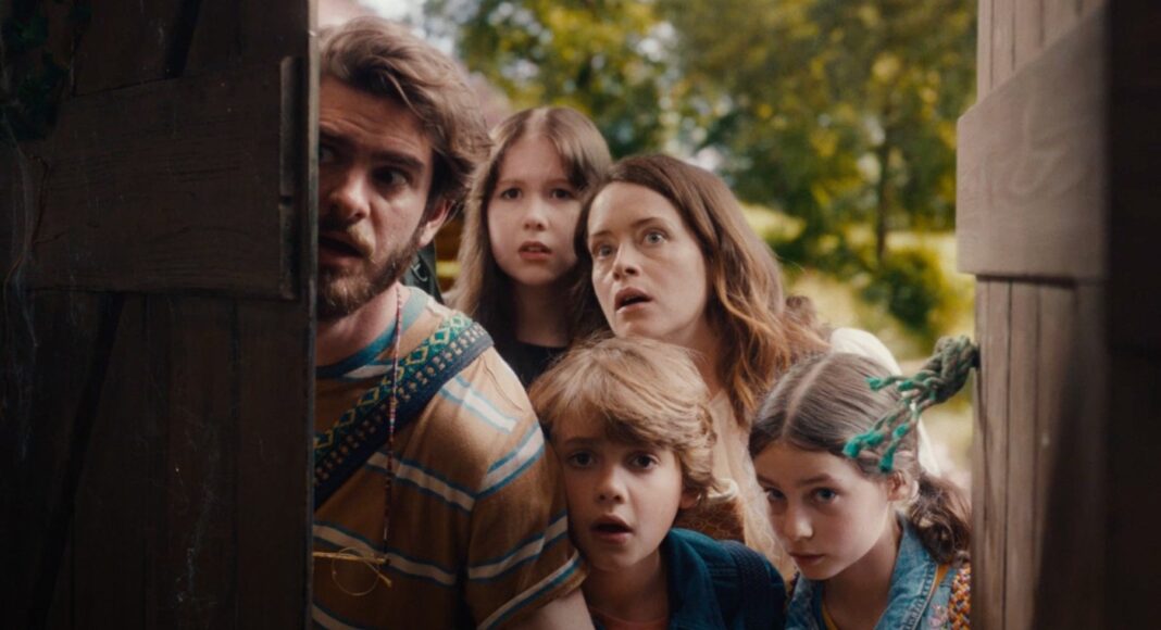 Five people—two adults and three children—peeking from behind a wooden door, looking startled in a park setting.