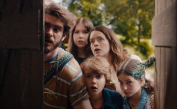Five people—two adults and three children—peeking from behind a wooden door, looking startled in a park setting.