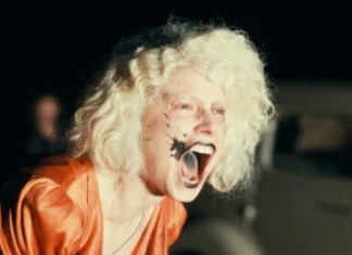 Person with a pale, curly wig and orange clothing screaming with mouth open, black makeup on the cheek, in a dark outdoor setting near a van.