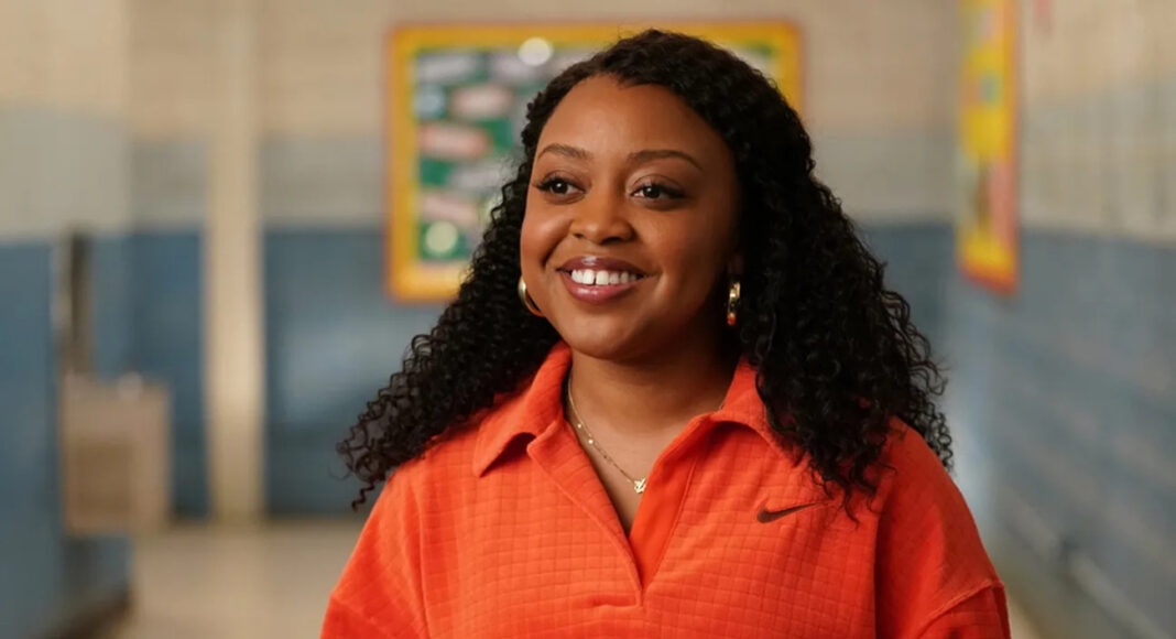 Smiling woman with curly hair wearing an orange polo in a school hallway? portrait in a soft focus setting.