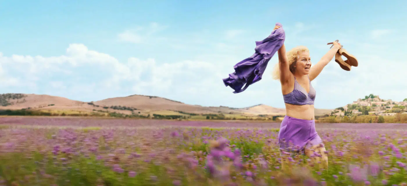 Woman in a purple outfit dancing in a lavender field, arms raised with a scarf and a hat in hand under a blue sky