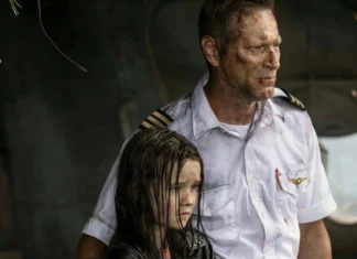 Wounded pilot in a torn white uniform stands beside a young girl with wet, smoky hair in a ruined aircraft cabin, both looking off to the side.