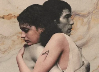 Two people embrace back-to-back in a muted, painterly scene; one arm shows a visible A tattoo on the upper arm.