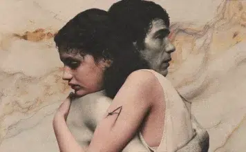Two people embrace back-to-back in a muted, painterly scene; one arm shows a visible A tattoo on the upper arm.