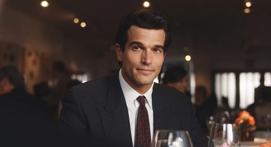 american horror story Man in a dark suit and tie sits at a dining table in a fine-dining restaurant with wine glasses nearby.