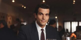 Man in a dark suit and tie sits at a dining table in a fine-dining restaurant with wine glasses nearby.