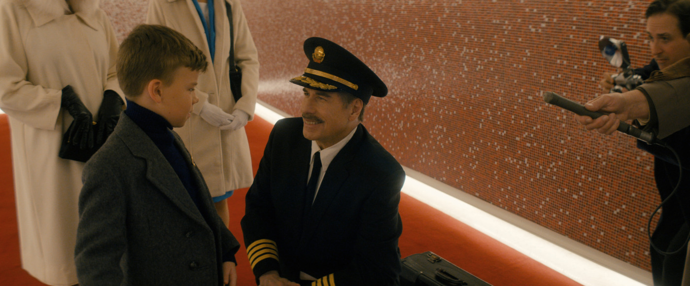 Pilot in uniform kneels to talk with a young boy in a gray coat on a red carpet. Mosaic wall in the background.