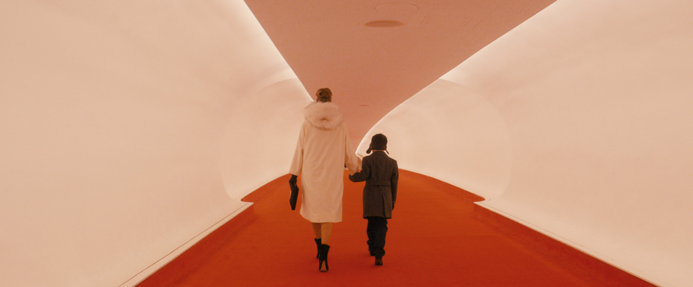 Adult and child walk hand in hand down a red carpeted, futuristic-looking hallway with curved white walls.