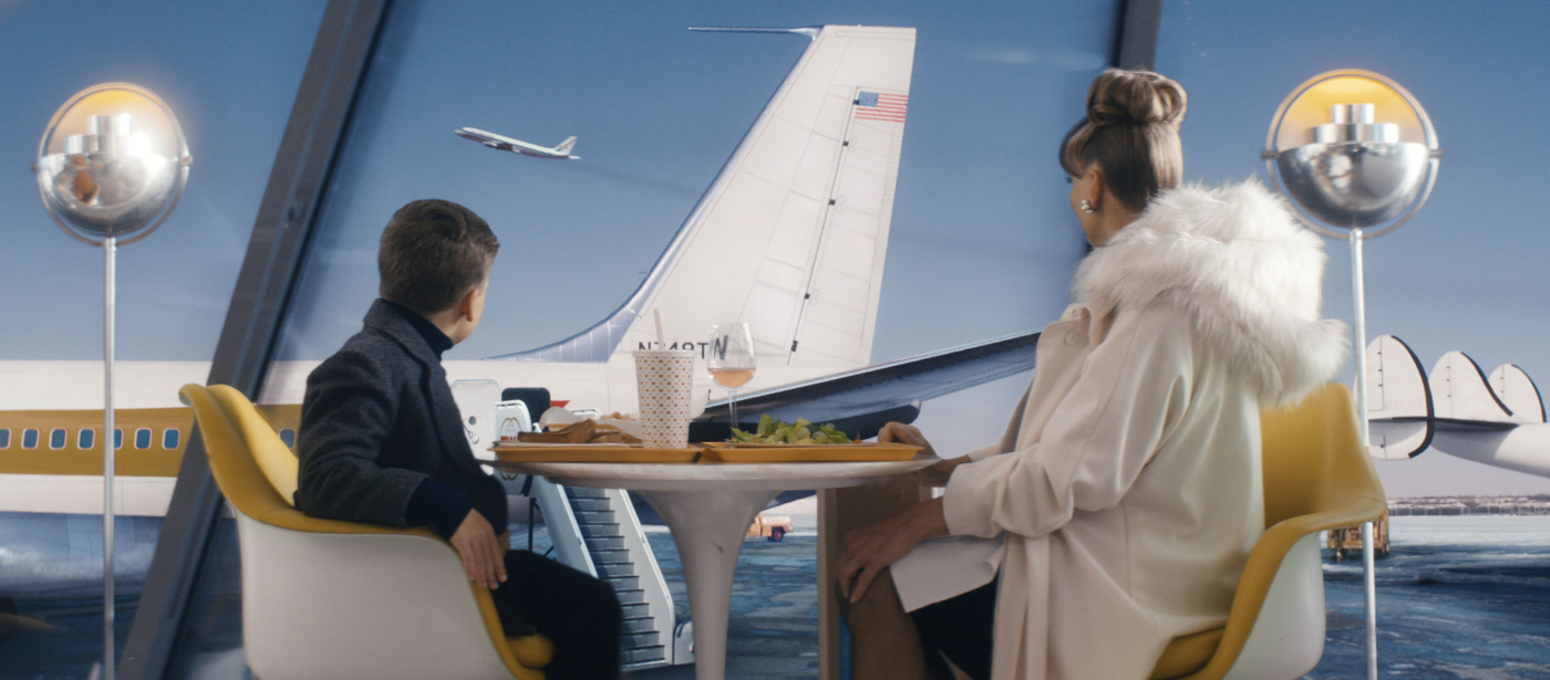 Couple dining at a round table in a bright airport lounge, with a large window view of a plane outside behind them.