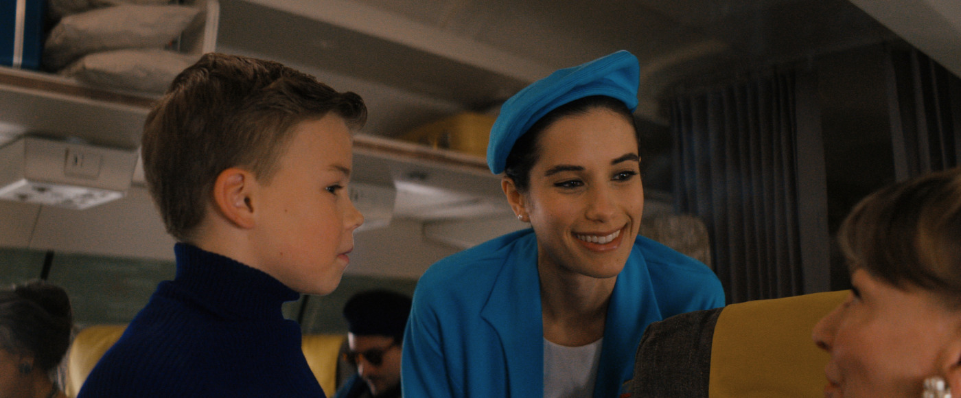 Flight attendant in blue uniform leaning toward a smiling boy in a blue sweater on an airplane interior.