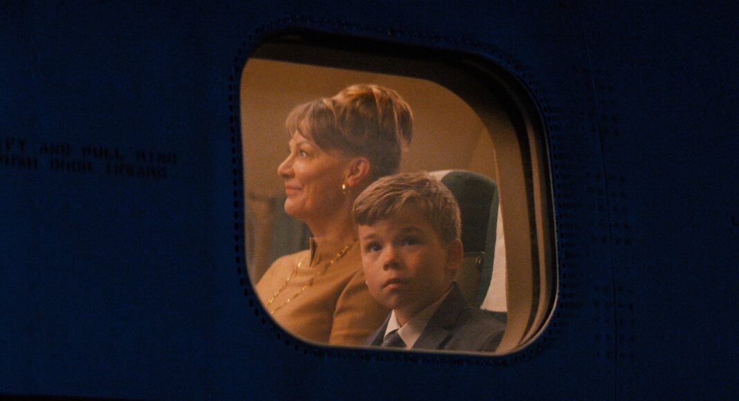 Woman with short hair and a young boy in a suit seated by an airplane window, illuminated by cabin light, looking ahead.